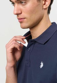 Young man adjusting the collar of a navy blue polo shirt with a white embroidered logo on the chest.
