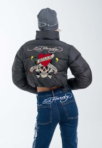 Schwarze Daunenjacke mit besticktem Schädel- und Herzdesign, Text "KILLS" und "LOVE SLOWLY", kombiniert mit blauen Jeans mit ähnlicher Bestickung.