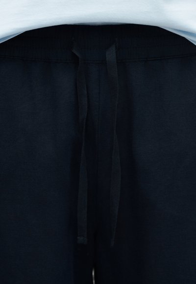 Κοντινό πλάνο στη μέση μαύρου παντελονιού φόρμας με μαύρα κορδόνια και μια ματιά σε ένα λευκό πουκάμισο από πάνω.