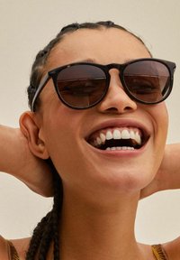 Occhiali da sole neri con lenti circolari, una montatura sottile e vetri scuri. Lo sfondo è neutro, mettendo in risalto il design del prodotto.