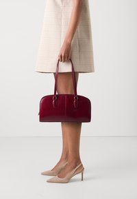 Burgundská kožená kabelka zaobleného tvaru, s dvojitými horními uchy a hladkým povrchem. V kombinaci s lehkou kostkovanou sukní a béžovými podpatky.