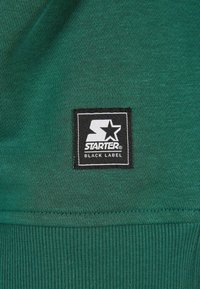Zeleni materijal s tkanim uzorkom, s crnom etiketom na kojoj piše "STARTER BLACK LABEL" i bijelim logotipom zvijezde.