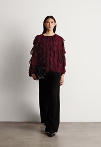 Burgundy ruffled blouse with sheer layers, paired with black wide-leg trousers and a black clutch. Minimalistic background setting.