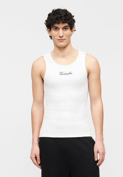 Jeune homme portant un débardeur blanc avec le texte « Endorphin », associé à un pantalon noir, debout devant un fond clair uni.