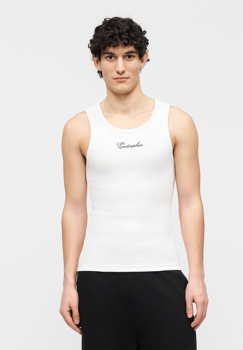 Jeune homme portant un débardeur blanc avec le texte « Endorphin », associé à un pantalon noir, debout devant un fond clair uni.