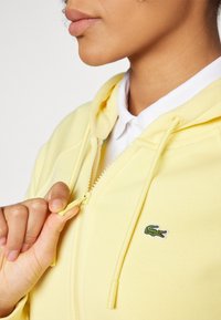 Geltona zip-up gobtuvė su kengūros kišene, su žaliu siuvinėtu krokodilo logotipu ir traukimo virvutėmis su plastikiniu antgaliu.