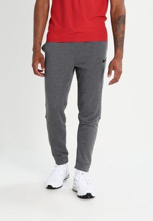 Grijze Sweatpants met een taps toelopend ontwerp, met een zwart Nike-logo. Het model draagt witte sportschoenen, op een neutrale achtergrond.
