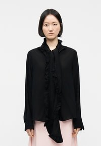 Mujer con cabello negro liso en corte bob, vestida con una blusa negra con volantes y mangas largas, y una falda rosa pálido, de pie frente a un fondo liso.