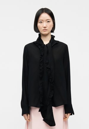 Femme aux cheveux noirs coupés au carré et raides, vêtue d'un chemisier noir à volants et manches longues, et d'une jupe rose pâle, se tenant devant un fond uni.