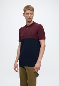 Polo shirt featuring a burgundy upper section and navy lower section, made of cotton, structured collar, three-button placket, and subtle logo.