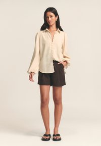 Cream blouse with long puff sleeves and button front, paired with dark brown shorts and black sandals. Smooth texture, loose fit design.