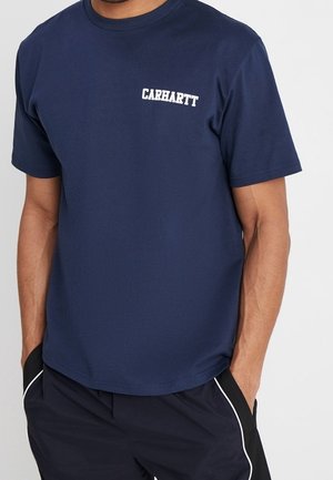 Mand iført en mørkeblå kortærmet t-shirt med teksten "CARHARTT" på brystet, hænderne i lommerne på sorte bukser med hvid piping.