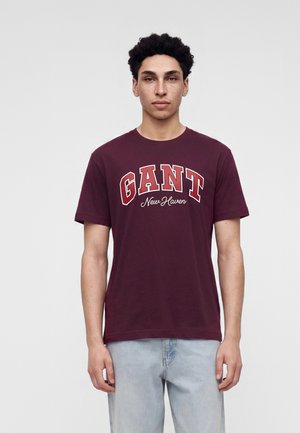 Jeune homme aux cheveux bouclés portant un t-shirt bordeaux GANT New Haven et un jean bleu clair, debout devant un fond blanc uni.