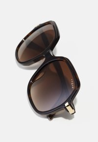 Ochelari de soare supradimensionați, pătrați, cu cadru din fâcă, lentile maro cu degradé și accesorii aurii pe care este gravat numele brandului "vogue."