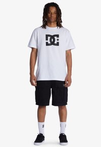 Weißes Baumwoll-T-Shirt mit großem schwarzem DC-Logo, kombiniert mit schwarzen Cargoshorts mit Taschen. Schwarze Sneaker und weiße Socken mit Logo.