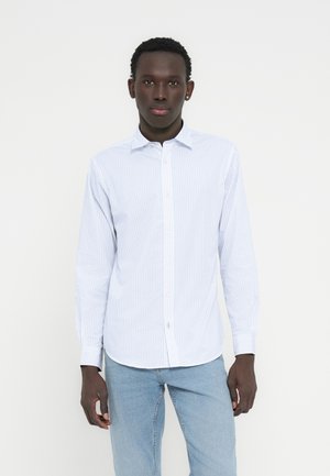 Hombre de pie mirando hacia adelante con una camisa blanca con botones y diseño, y vaqueros azul claro, sobre un fondo blanco liso.