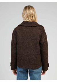 Femme blonde portant une veste en laine texturée marron foncé et un jean bleu, se tenant de dos contre un fond clair.