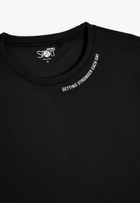Camiseta deportiva negra de tejido suave y ligero. Presenta un cuello redondo y texto blanco "MEJORANDO CADA DÍA" en el cuello.