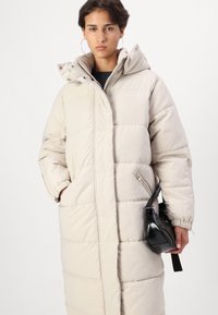 Manteau long rembourré en beige clair avec un col haut, fermeture éclair et boutons-pression. Tissu texturé et poches latérales. Sac à main noir avec bandoulière.