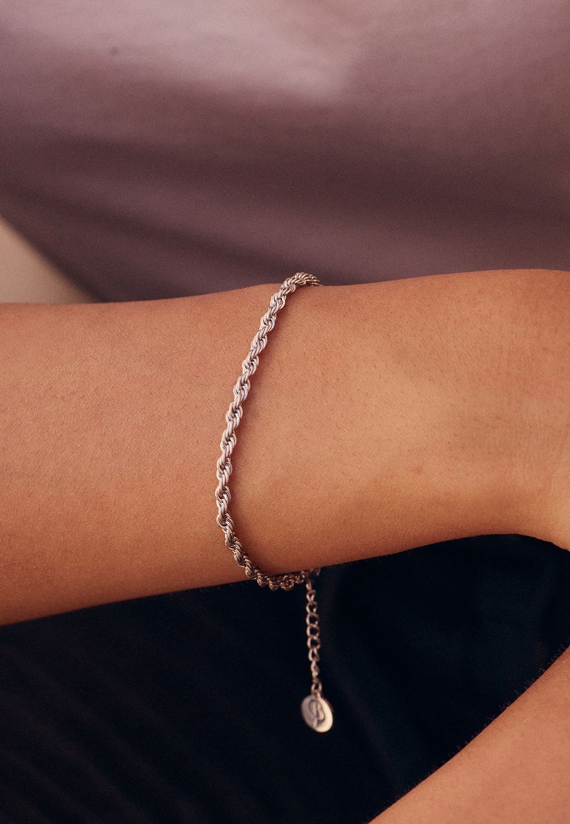 Srebrna spirala zapestnica na zapestju z visečo verigo in majhnim okroglim obeskom; gladka tekstura in poliran zaključek.