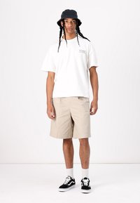 Tricou alb din bumbac cu detalii grafice, pantaloni scurți bej, clopot negru și adidași negri cu accente albe. Stând în fața unui fundal simplu.