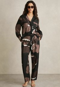 Crni i smeđi uzorak loungewear seta s košuljom s kragnom na dugmad i hlačama s elastičnim pojasom, u kombinaciji s tamnim naočalama i sandalama s trakicama.