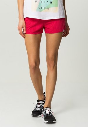 Vrouw draagt felroze sportshorts, een wit T-shirt met opdruk en zwarte hardloopschoenen met witte veters, staand tegen een effen achtergrond.