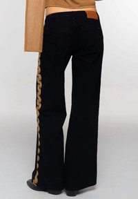 Vaqueros acampanados negros con un patrón de cadena dorada a lo largo de las costuras laterales y detalles de parches en la parte trasera, hechos de tela vaquera.