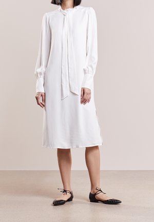 Femme portant une robe midi blanche à manches longues avec un détail nœud au col et des ballerines noires pointues avec liens à la cheville, debout sur un sol neutre.