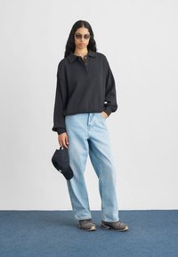 Svart ribbad tröja med krage, blå baggy jeans, grå skor och svart keps. Modellen bär solglasögon; bakgrunden är helt vit.