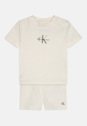 Conjunto de algodão em duas peças, apresentando uma parte de cima de manga curta em creme com um logótipo e calções combinando com um logótipo. Textura suave, design simples.