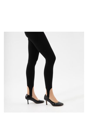 Jambes portant des leggings noirs à brides associées à des chaussures à talons hauts en cuir verni noir sur fond blanc.