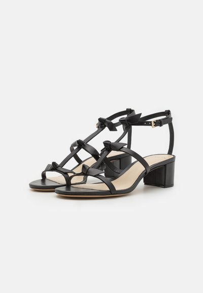 Svarta klacksandaler med flera remmar, som har rosetter, rund tå och blockklack. Slät läder och beige innersula synlig.