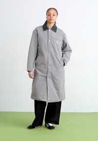 Cappotto lungo grigio con bottoni e colletto verde, dotato di taschino e tasche laterali, indossato sopra pantaloni neri e sneakers.