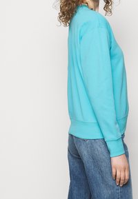 Tyrkysová mikina s kulatým výstřihem, dlouhými rukávy a vroubkovanými manžetami, spárovaná s volnými modrými džíny, zachycena z boku.