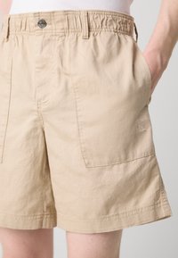 Béžové šortky vyrobené z lehkého materiálu s elastickým pasem, vybavené bočními kapsami a jemně vyšitým logem na přední straně.