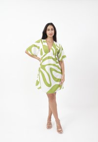 Weißes Kleid mit grünen abstrakten Mustern, V-Ausschnitt, kurzen weiten Ärmeln und einem lockeren Schnitt. Kombiniert mit klaren Sandalen mit Absatz.