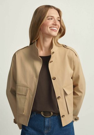 Uśmiechnięta kobieta o długich blond włosach, ubrana w beżową, zapinaną na guziki kurtkę, czarną koszulę, niebieskie dżinsy oraz czarny pasek ze złotą klamrą.