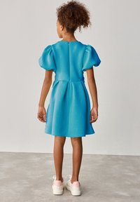 Blaues Kleid mit kurzen Puffärmeln, strukturiertem Stoff mit geprägten Buchstaben und Schleifen, ausgestellter Rock, kombiniert mit weißen Sneakers mit rosa Akzenten.