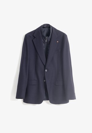 Blazer navy con tejido texturizado, solapas de pico, dos bolsillos delanteros y un forro interno con cremallera. Presenta botones de acento marrón.