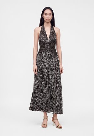 Femme portant une robe noire sans manches à col halter avec des pois blancs et des sandales dorées à talons hauts, debout devant un fond uni.