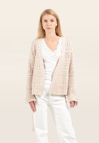 Beige gestrickte Strickjacke mit offenem Vorderteil, langen Ärmeln und großen Häkelmustern. Getragen über einem weißen Oberteil und kombiniert mit weißen Hosen.