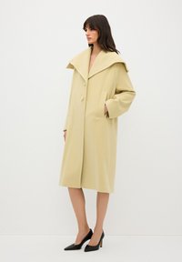 Beige oversized uldfrakke med en bred krave, to forlommer og to knapper. Skræddersyet pasform, knælang, glat tekstur, båret over sorte hæle.