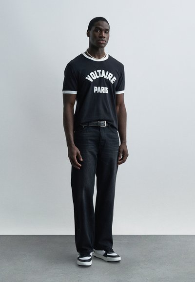 Νεαρός άντρας που στέκεται μπροστά σε γκρι τοίχο, φορώντας μαύρο μπλουζάκι "Voltaire Paris", σκούρο τζιν, μαύρα και λευκά αθλητικά παπούτσια και περιδέραιο με μαργαριτάρια.