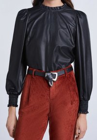 Svart läderblus med puffärmar och volangkrage; kombinerad med rödbruna kordbyxor med texturerad yta och bälte i midjan.