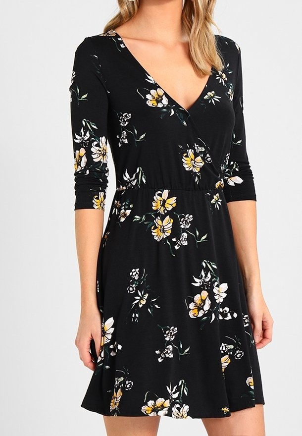 Femme portant une robe portefeuille noire à fleurs jaunes et blanches avec des manches trois-quarts, debout devant un fond uni.