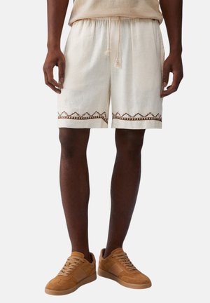 Uomo che indossa pantaloncini beige con coulisse e rifinitura geometrica marrone, sneakers in camoscio color cuoio, in piedi contro uno sfondo uniforme.