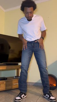 Jeans de ganga azul com corte reto, usados com uma t-shirt branca e ténis pretos. Fundo neutro com uma televisão e uma lareira.