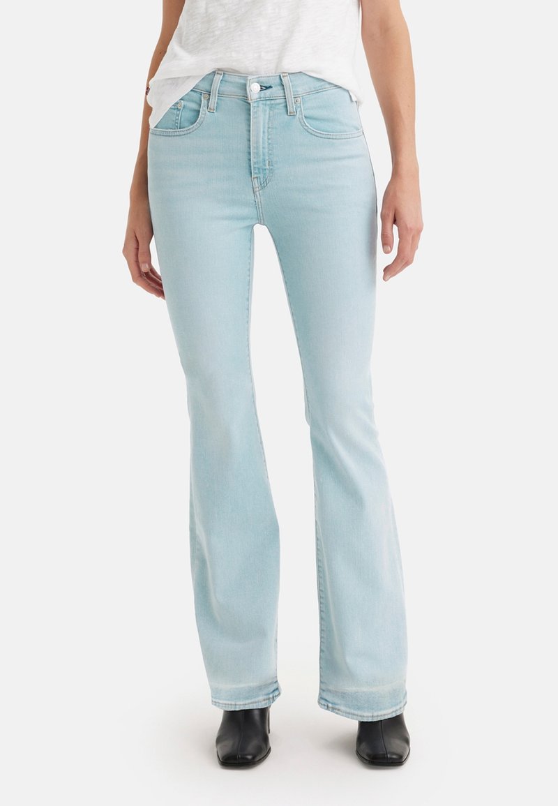Jean flare bleu clair en denim, avec une taille haute, cinq poches et des détails de couture subtils. Le modèle porte des bottines noires.