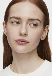 Złote kolczyki w kształcie niedźwiedzia, o gładkiej fakturze i wypolerowanej powierzchni, noszone przez modelkę z prostymi, średniej długości włosami.